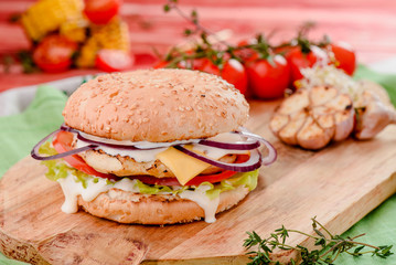 burger with chicken cutlet, tomatoes, cheese, lettuce and red sauce on a wooden board on a red wooden background, decorated with napkins, chili pepper and cherry tomatoes. close up