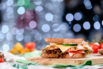 New Year's sandwich with chicken, tomatoes, cheese, sauce and lettuce on a wooden board. Against the background of a blurred blue Gerland. close-up
