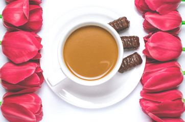 White coffee cup stands on a white plate with white background near multi-colored tulips