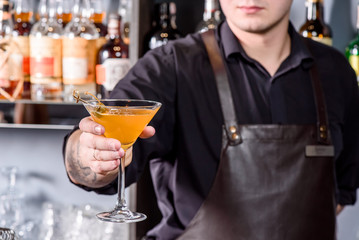 Barman in brown leather apron holds glass of orange cocktail decorated with capers at bar counter background. Glass of orange cocktail with vodka, syrup and wine at bar background. Close up. Space