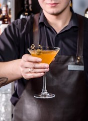 Barman in brown leather apron holds glass of orange cocktail decorated with capers at bar counter background. Glass of orange cocktail with vodka, syrup and wine at bar background. Close up. Space