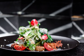 Caesar's creative feed. Traditional salad with grilled chicken, tomatoes, cheese, spinach and cream sauce on a dark plate close-up. Warm salad with meat, vegetables and spinach on a black plate