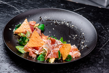 salad with prosciutto, nachos, tomatoes, cheese and spinach on a black matte plate close-up on a dark background. close-up. space. photo series