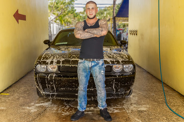 A proud young man stands in front of his soapy car. The masculine man with tattoos takes a moment...