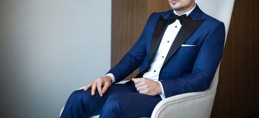 Man model in expensive custom tailored blue tuxedo, suit sitting and posing indoors