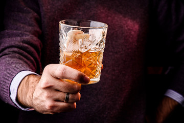Man in a purple sweater holding a beautiful faceted glass of whiskey decorated with mushrooms. Conceptual supply of whiskey. dark atmosphere. close up