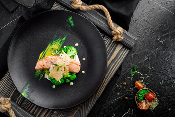 Beautiful serving of baked salmon with creamy sauce, with broccoli, tomatoes on a black matte plate on a rustic wooden tray on black background. Beautiful composition on a dark background. Top view