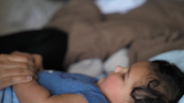 A mixed race African American mom pokes her chubby baby on the cheek and kisses him as he lays on his back in the bed with a fluffy brown comforter in the background.