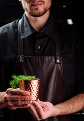 Man in a dark leather apron holds cocktail in a copper glass decorated with dill. Close up. dark atmosphere.