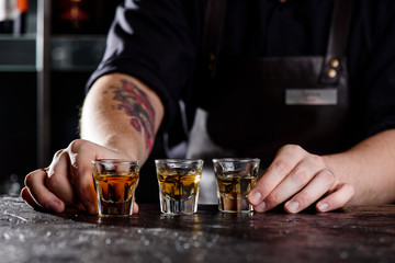 Bartender in a dark leather apron puts three glasses of whiskey on a dark wooden bar in a nightclub. Close-up. Spa ce