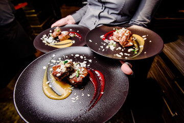 The girl waiter carries three plates of food. Meat steak garnished with two sauces on a dark plate. Close-up. Wide angle photography. 