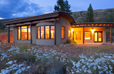 Modern, sustainable, energy efficient earth berm house home with fire resistant stucco exterior and xeriscape drought tolerant native plants landscaping with lights on at dusk twilight