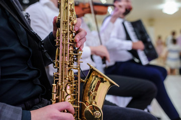 close up photo of a man in black suit that plays a saxophone near a violin and a accordion musicians