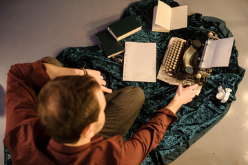 top view of man with a glass of wine in his hands sitting near a vintage typewriter on a piece of newspaper and dark blue velvet fabric on the floor with sheets of paper and books arround