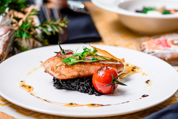 Beautiful serving of roasted salmon fillet and risotto with cuttlefish ink on a white plate with a signature sauce and Cherry tomatoes in a restaurant. close up