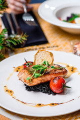 Beautiful serving of roasted salmon fillet and risotto with cuttlefish ink on a white plate with a signature sauce and Cherry tomatoes in a restaurant. close up