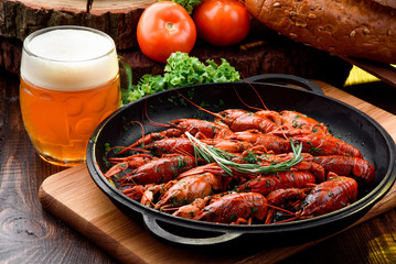 Beer Snack. Light beer in a transparent bobkale and Boiled crayfish in a cast-iron pan on a light wooden board on a dark wooden table, decorated with vegetables. Close-up. Space