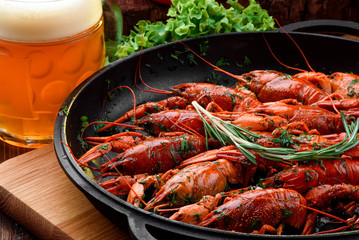 Beer Snack. Light beer in a transparent bobkale and Boiled crayfish in a cast-iron pan on a light wooden board on a dark wooden table, decorated with vegetables. Close-up. Space