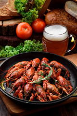 Beer Snack. Light beer in a transparent bobkale and Boiled crayfish in a cast-iron pan on a light wooden board on a dark wooden table, decorated with vegetables. Close-up. Space