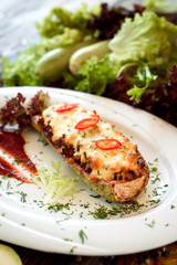 Baked stuffed zucchini with cheese and chilli on a white plate with red sauce. Wooden rustic background with vegetables and lettuce. Close-up. Space