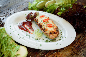Baked stuffed zucchini with cheese and chilli on a white plate with red sauce. Wooden rustic background with vegetables and lettuce. Close-up. Space