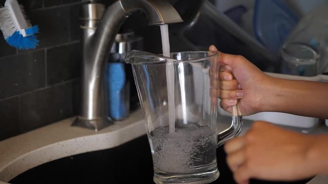 A Close Up Of An Mixed Ethnicity African American Woman Filling Tap Water Into A Glass Pitcher From The Kitchen Sink.