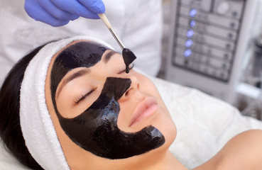 The procedure for applying a black mask to the face of a beautiful woman. Spa treatments and face care in the beauty salon.