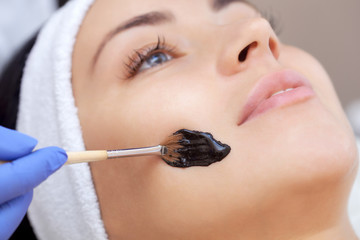 The procedure for applying a black mask to the face of a beautiful woman. Spa treatments and face care in the beauty salon.