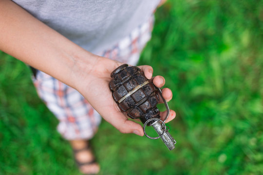 Closeup top view of white kid hand holding real old grenade isolated on green grass background. Horizontal color photography.