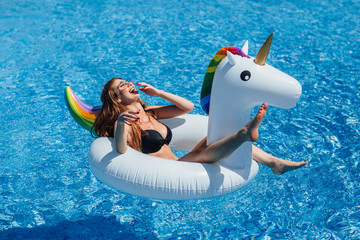 Young beautiful brown-haired girl with a good figure tan in the pool on an inflatable unicorn in a black swimsuit. © Тарас Нагирняк
