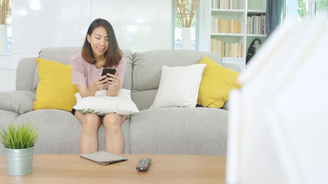 Young Asian woman using smartphone checking social media feeling happy smiling while lying on the sofa when relax in living room at home. Lifestyle latin and hispanic ethnicity women at house concept.