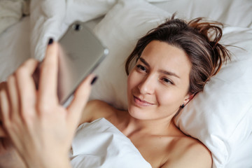 Obraz na płótnie Canvas Beautiful girl lying on the bed, smiling and looking into the phone