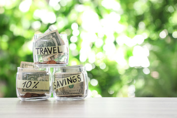 Glass jars with money for different needs on table against blurred background