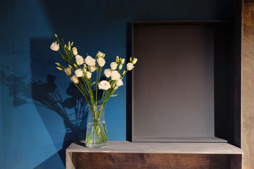 A bouquet of white roses in a glass vase stands against a dark blue wall. Wooden shelves.