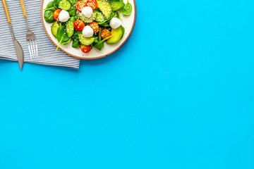 Fresh salad with mozzarella, spinach, cherry tomatoes, cucumber on plate on blue background top view space for text