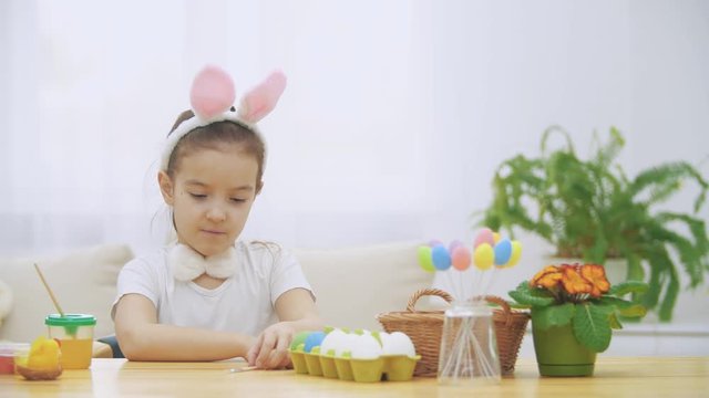 Little cute girl is having fun holding a paint-brush in her left hand. Girl with a beauty spots at her face watching at nude paint-brushes, sitting at the wooden table with Easter decorations. Girl