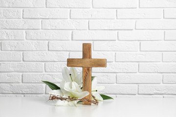 Wooden cross, crown of thorns and blossom lilies on table against brick wall