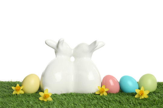 Ceramic Easter bunnies and dyed eggs on green grass against white background