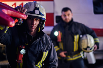 Two firemen wearing a protective uniform standing next to a fire truck. Arrival on call at night...