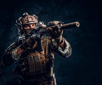 Elite unit, special forces soldier in camouflage uniform holding assault rifle and aiming with optical sight. Studio photo against a dark textured wall