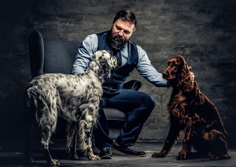 Middle-aged hunter dressed in elegant clothes sits on a sofa with his two purebred Setter dogs....