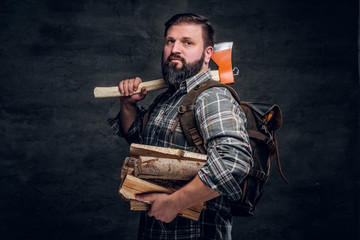Portrait of a bearded woodcutter with a backpack dressed in a plaid shirt holding firewood and ax....