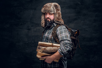 Portrait of a bearded woodcutter with a backpack dressed in a plaid shirt and trapper hat holding...