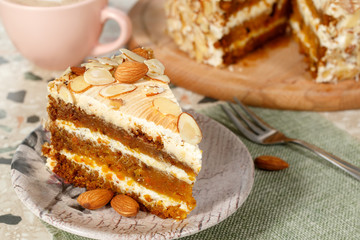 Close-up the slice  of vegetarian carrot cake with almonds on a saucer and a fork near. Pink cup of coffe and wooden board with cake in the background.