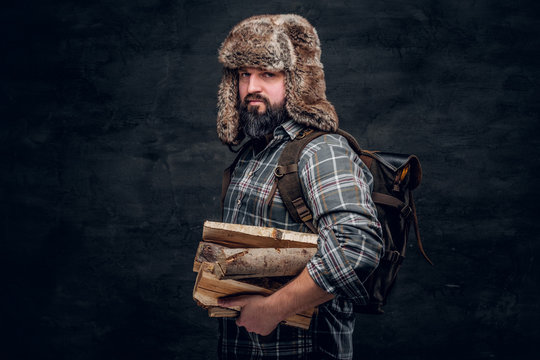 Portrait of a bearded woodcutter with a backpack dressed in a plaid shirt and trapper hat holding firewood. Studio photo against a dark textured wall