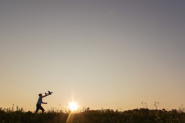 Black silhouette of cute happy cheerful child running fastly along grassy hill at countryside holding big toy plane in hand. Boy playing during sunset time in evening. Horizontal color photography.