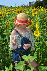 Mädchen mit Strohhut steht im Sonnenblumen Feld und schaut nach unten, Hände vor dem Körper