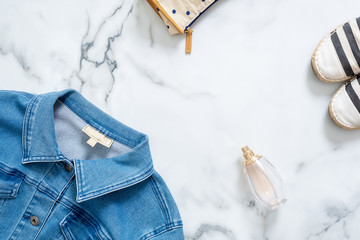 Desk of beauty blogger with feminine jeans jacket, cosmetic bag, fragrance bottle of perfume, fashionable striped sandals. Retro fashioned women's clothing and accessories on marble background.