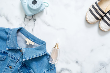 Beauty blogger desk with feminine jeans jacket, hipster instant photo camera,  fragrant bottle of perfume, fashionable striped sandals. Casual women's clothing on marble background on flat lay style