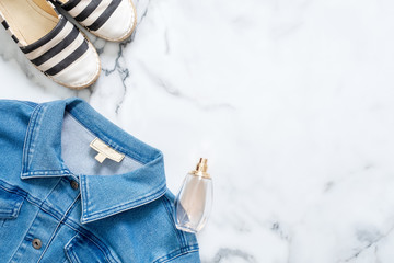Jeans jacket, bottle of perfume, striped summer sandals on marble background. Flat lay design composition with retro fashioned feminine clothing and accessories. Beauty blogger lifestyle.